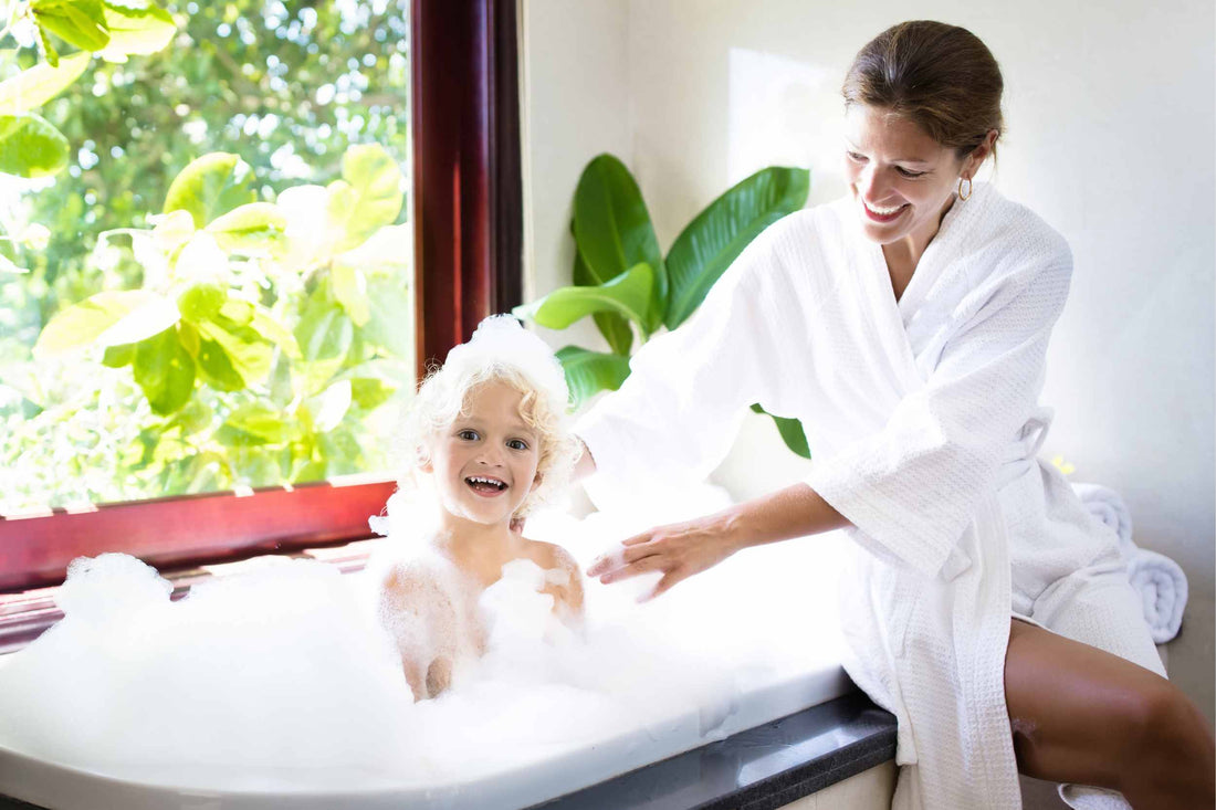 Toddler enjoying having hair washed in a soapy bathtub. His mother is in a white towel robe sat on the edge of the bath smiling at him. There is a lot of greenery in the background.
